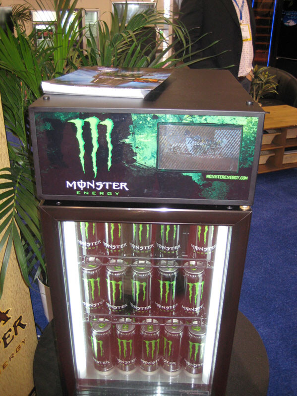 A small glass-door cooler stocked with Monster Energy cans, topped by a Monster Energy panel with a small video screen. A brochure sits on top, with palm plants nearby.