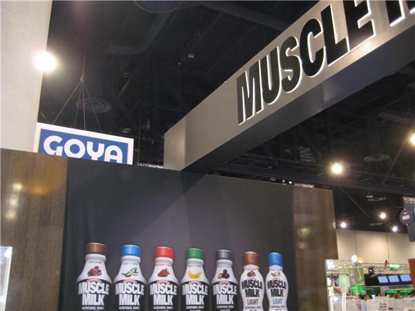A booth wall displays a row of Muscle Milk bottles below a large Muscle Milk sign; a Goya sign appears in the background of the convention hall.