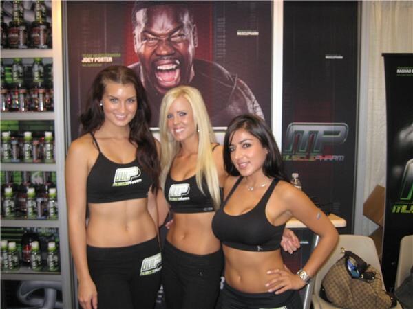 Three smiling women in black MusclePharm crop tops and pants pose together in front of a booth with shelves of bottles and a poster of a shouting man.