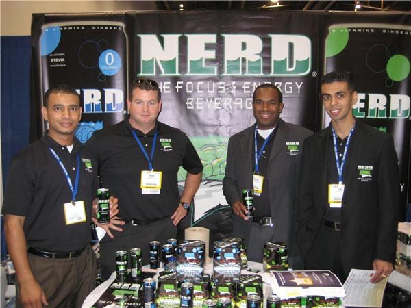 Four men in black shirts or jackets with lanyards stand at the NERD booth, holding cans, with NERD banners behind them and product cans and boxes on the table.