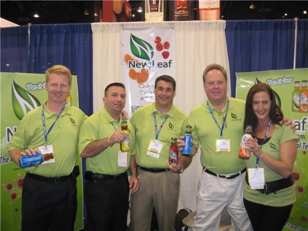 Four men and a woman in green New Leaf polo shirts and lanyards stand together at the booth, holding bottles of tea, with New Leaf banners behind them.