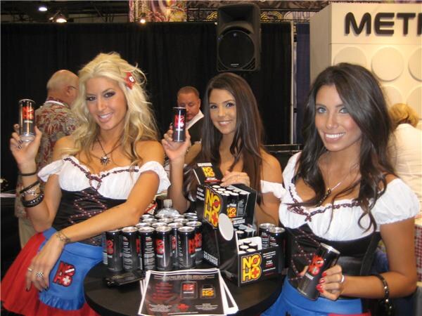 Three women in white peasant tops and dirndl-style outfits hold black NOHO cans, with a table of NOHO cans and display boxes in front of them.