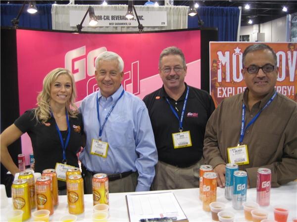 A woman and three men with lanyards stand behind a table of GoGirl cans and sample cups under a pink GoGirl banner and a Nor-Cal Beverage Co., Inc. sign.
