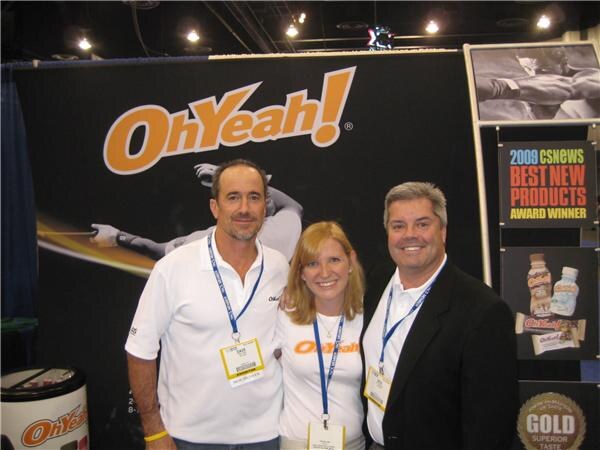Two men and a woman in lanyards smile together in front of an OhYeah! banner, with a CSNews Best New Products Award Winner sign and product images beside them.