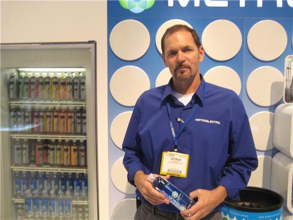 Patrick, wearing a blue Metroelectro shirt and an exhibitor badge, holds a bottled water in front of a lit cooler of bottled drinks and a blue wall with white circles.