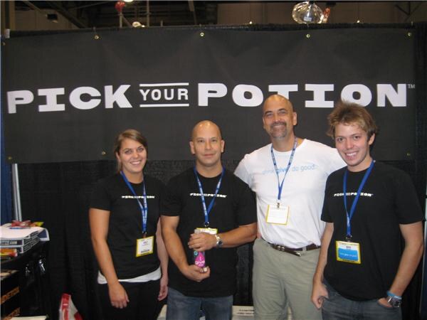 Two men and a woman in black shirts and a man in a white shirt, all with lanyards, stand in front of a black Pick Your Potion banner. One man holds a small bottle.