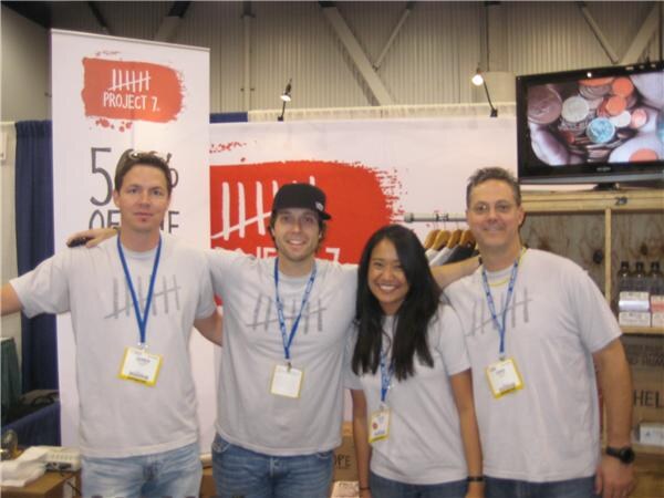 Three men and a woman in gray Project 7 tally-mark t-shirts and lanyards stand arm in arm at the booth, with Project 7 banners and a screen showing coins behind them.