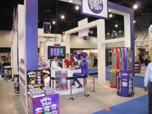 Purple Stuff booth with white and purple frame structure, a person in a purple bodysuit and wig seated on a stool, two staff at the counter, and shelves and displays of Purple Stuff cans.