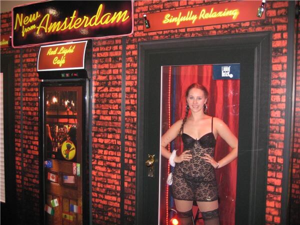 A woman in black lace lingerie stands in a doorway with a red curtain beside a brick wall with neon signs reading "New from Amsterdam," "Red Light Café" and "Sinfully Relaxing."