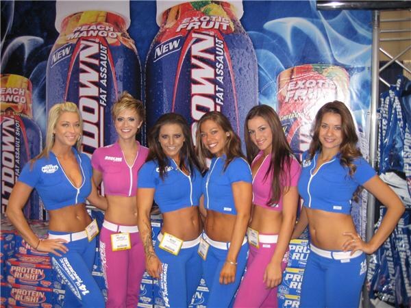 Six women in matching blue and pink outfits with badges pose in front of large Redline can graphics and stacked Protein Rush cases.