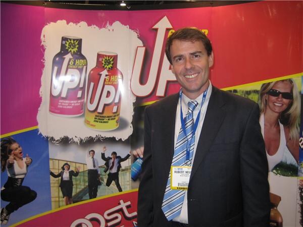 Robert Neu, in a dark suit and striped tie with an exhibitor badge, smiles in front of a pink UP! backdrop showing UP! energy shot bottles.