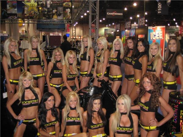 A large group of women in black and yellow Rockstar Energy Drink outfits pose around a motorcycle, with CardTronics and Icee signs in the background.