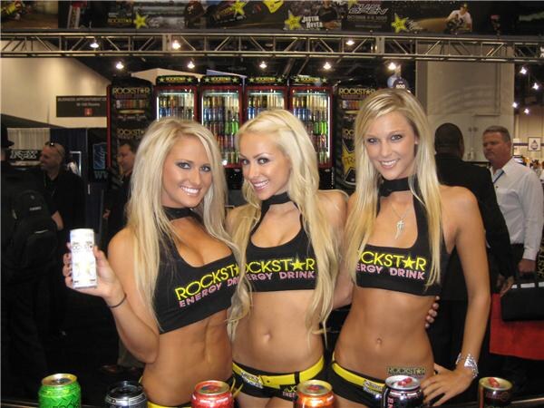 Three smiling women in Rockstar Energy Drink tops stand at a counter with cans in front, one holding a white can, before Rockstar refrigerated coolers.