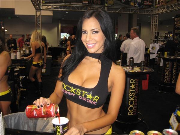 A smiling woman in a Rockstar Energy Drink top pours a red can into a cup at a booth with Rockstar branded tables behind her.