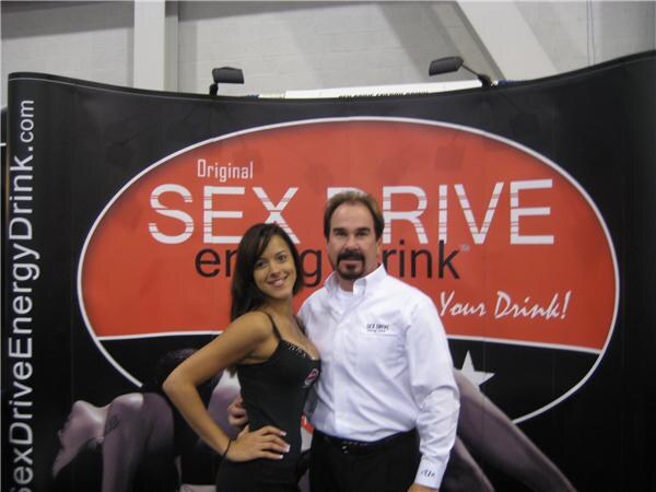 A man in a white shirt and a woman in a black dress stand together in front of a black Sex Drive Energy Drink backdrop with a red oval logo.