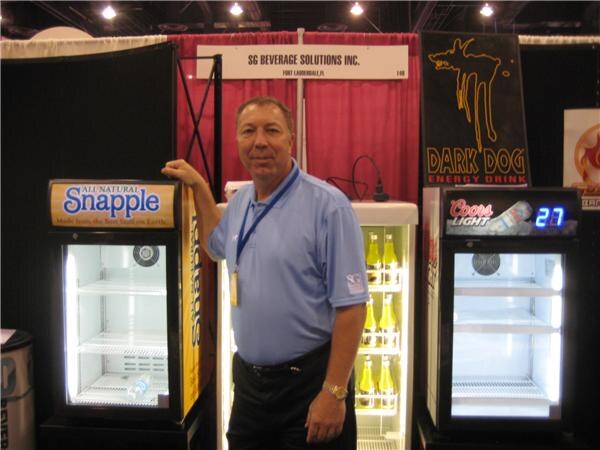 A man in a blue polo stands among lit coolers branded Snapple and Coors Light, near a Dark Dog Energy Drink banner and an SG Beverage Solutions Inc. sign.