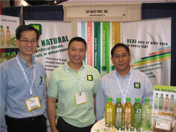 Three men with lanyards stand in front of green aloe vera banners at the SPI West Port, Inc. booth, with bottles of aloe drink on a table.