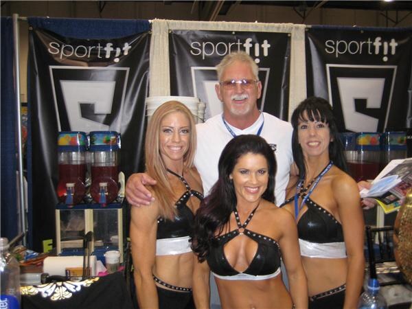 A man with sunglasses and a mustache stands with his arms around three smiling women in black studded tops in front of black Sportfit banners and two slush dispensers.