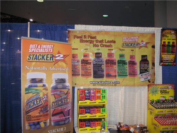 Booth display with Stacker 2 banners showing supplement bottles and energy shots, plus shelf displays of Stacker 2 shots. No people are visible.