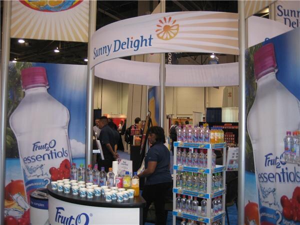 Sunny Delight booth with a curved overhead sign, Fruit2O Essentials bottle graphics, a table of sample cups, a shelf of bottled drinks and several people standing behind.