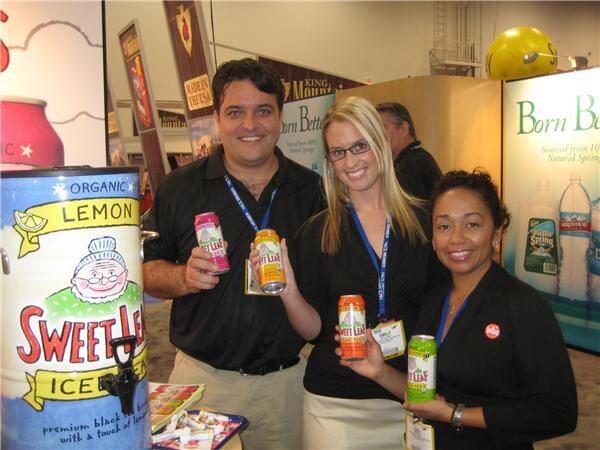 A man and two women in dark clothing and lanyards hold cans of Sweet Leaf tea beside a Sweet Leaf Lemon iced tea dispenser, with a Born Better water display behind them.