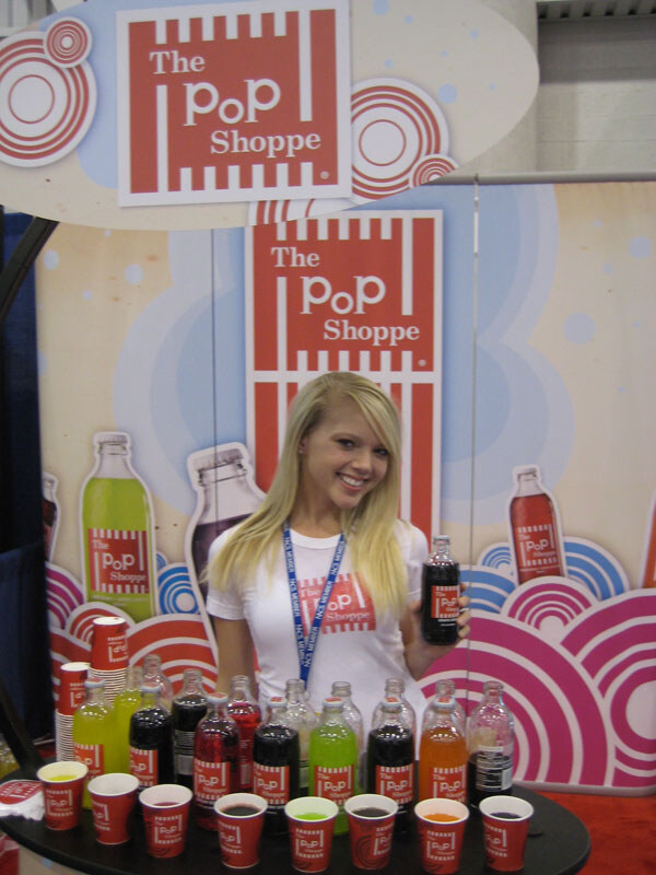 A smiling blonde woman in a white The Pop Shoppe shirt holds a bottle of dark soda behind a table of bottled sodas and red sample cups, in front of red and pink Pop Shoppe graphics.