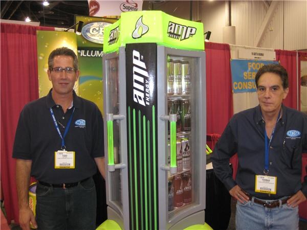 Todd, left, and Thomas, right, wearing CRD shirts and exhibitor badges, stand on either side of a tall green and black AMP Energy lighted cooler stocked with cans in a booth with red curtains.