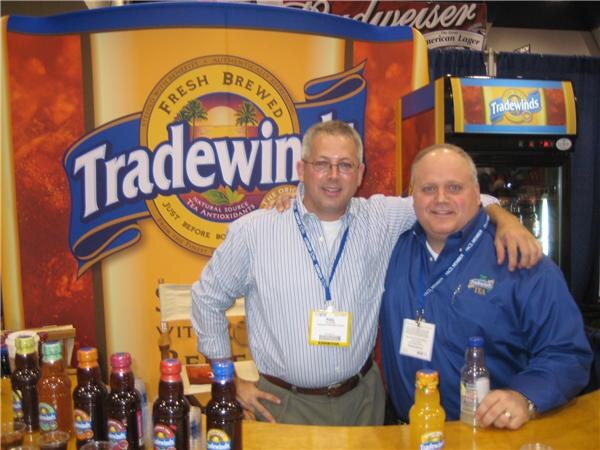 Two men with arms around each other stand at a counter in front of a large Tradewinds tea sign, with bottles of tea on the counter and a Tradewinds cooler behind them.