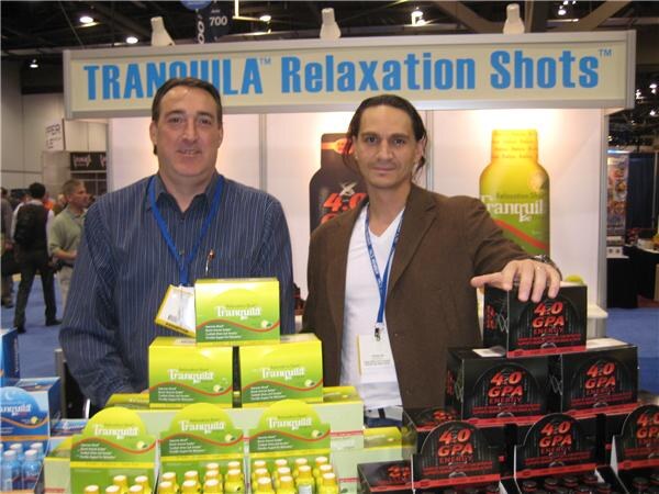 Two men with lanyards stand behind stacked boxes of Tranquila relaxation shots and 4.0 GPA Energy shots under a Tranquila Relaxation Shots sign.