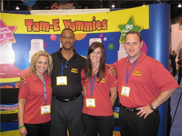 Four people in red and black shirts with name badges pose together in front of a Tum-E Yummies display showing bottles and a star reading 90 calories.