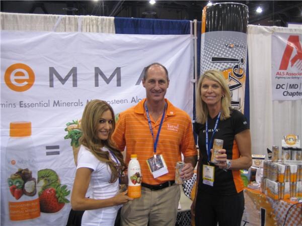 A man in an orange polo and two women stand together holding Vemma drink bottles and cans in front of a white banner reading Emma with fruit graphics.