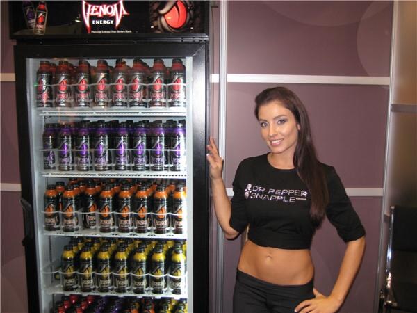 A woman in a black Dr Pepper Snapple top stands beside a lit cooler stocked with rows of Venom Energy drink bottles.