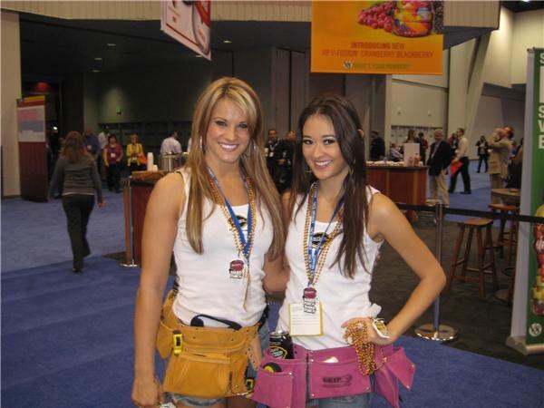 Two smiling women in white tank tops, lanyards and tool belts stand on a blue carpeted show floor below a banner introducing V-Fusion Cranberry Blackberry.