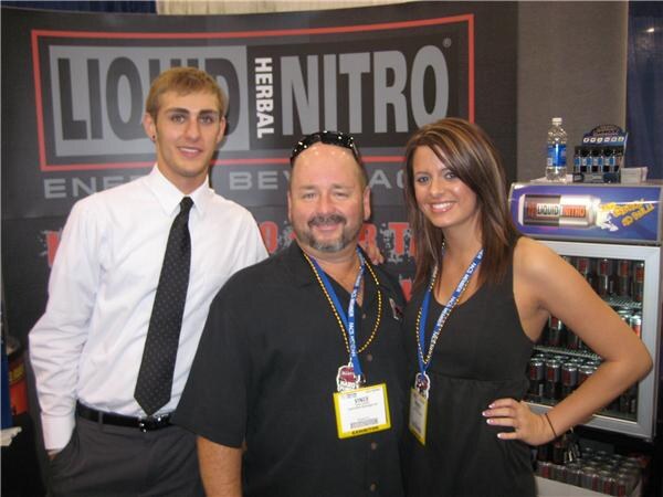 Vince, center, in a black shirt with sunglasses on his head, stands between a young man in a white shirt and tie and a woman in black, in front of a Liquid Herbal Nitro banner and cooler.