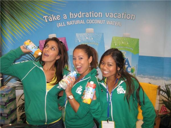 Three women in green Vita Coco jackets hold cartons of coconut water in front of a banner reading Take a hydration vacation, all natural coconut water.