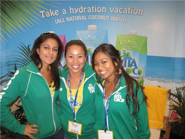 Three smiling women in green Vita Coco zip jackets and lanyards stand in front of a Vita Coco backdrop reading "Take a hydration vacation" with coconut water cartons and a beach scene.