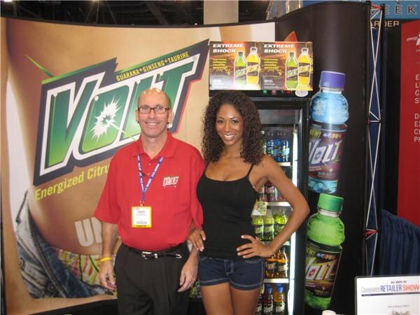 A man in a red Volt polo with glasses and a lanyard stands beside a woman in a black top and denim shorts at a Volt booth with a large logo banner and a lit beverage cooler.