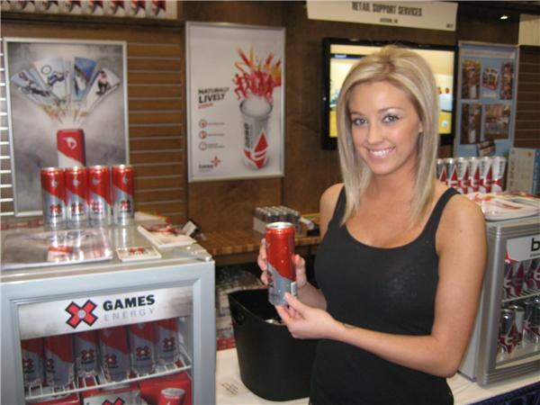 A smiling blonde woman in a black tank top holds a red X Games Energy can beside a small cooler stocked with the drink, with more cans displayed on shelves behind her.