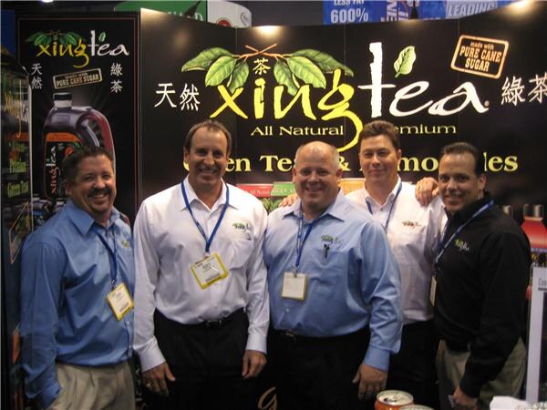 Five men in dress shirts and lanyards stand close together smiling in front of a black Xing Tea banner reading "All Natural Premium Green Tea & Lemonades" with a pure cane sugar badge.