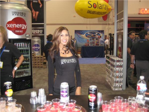 A woman in a black Xyience top stands behind a counter with cans and sample cups. A Xenergy can display, a cooler and a yellow Solaray blimp are behind her, with other people in the aisle.