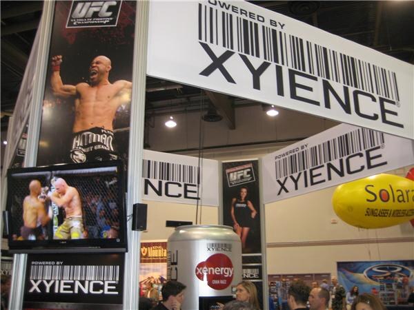 A Xyience booth with "Powered by Xyience" barcode signs, a UFC banner showing a shouting fighter, a video screen, a giant Xenergy can and a Solaray blimp, with attendees below.