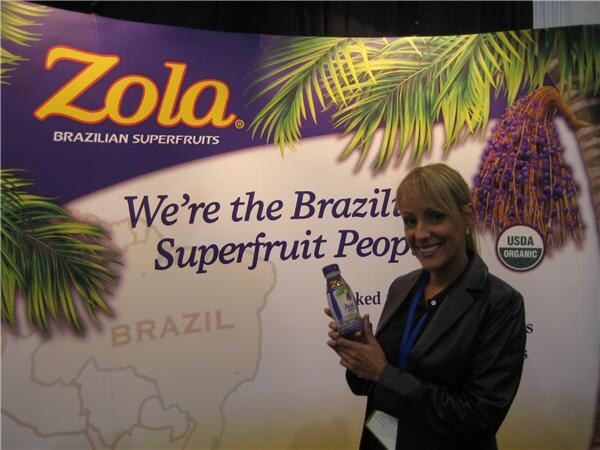 A smiling woman with blonde hair and a lanyard holds a bottle of Zola in front of a Zola Brazilian Superfruits backdrop reading "We're the Brazilian Superfruit People" with a USDA Organic seal.
