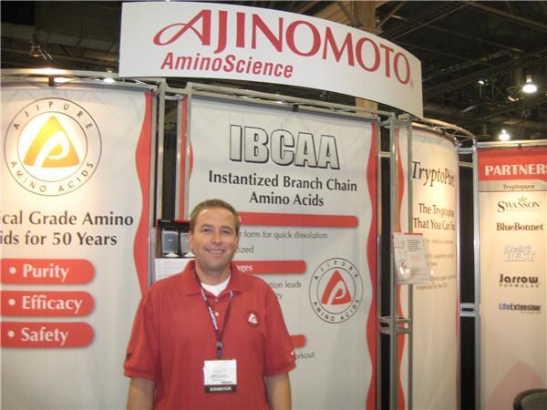A smiling man in a red polo with an exhibitor badge stands in the Ajinomoto AminoScience booth, in front of IBCAA and Ajipure panels and a partners sign listing Swanson, BlueBonnet, Jarrow Formulas and LifeExtension.