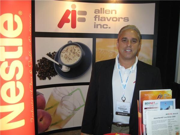 A man in a dark blazer with an exhibitor badge stands at the Allen Flavors Inc. booth beside a Nestlé banner, with BevNET.com printed materials on the counter in front of him.
