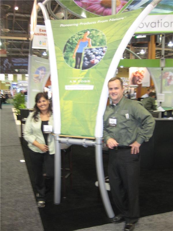 A woman in a light green jacket and a man in a green button-up shirt stand on either side of a curved green A.M. Todd banner on the show floor. The photo is slightly blurry.