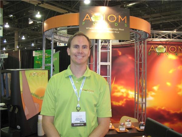 A smiling man in a green Axiom Foods polo with a lanyard and exhibitor badge stands in the Axiom Foods booth, with a sunset graphic backdrop, a beverage dispenser and small sample items on a table.