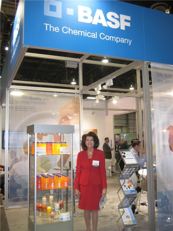 A woman in a red suit with a name badge stands in front of the BASF booth, which has a blue sign reading The Chemical Company and a glass case of orange and red bottled drinks.
