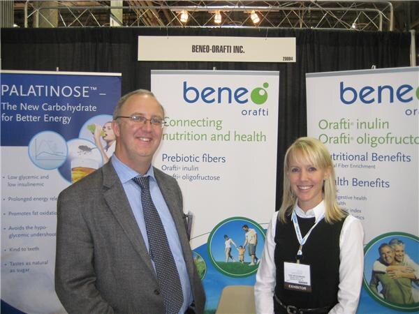 A man in a gray blazer and glasses and a blonde woman with an exhibitor badge stand in the Beneo-Orafti booth, in front of Beneo and Palatinose banners.