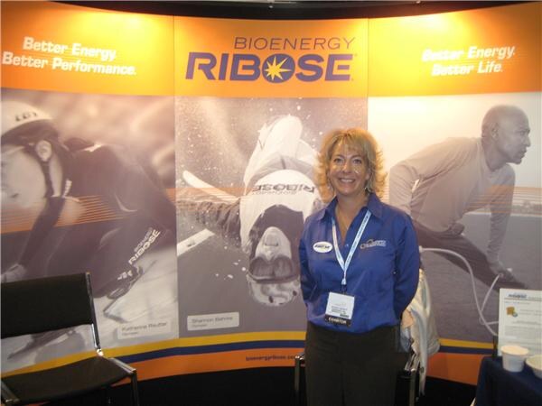 A smiling woman in a blue shirt with an exhibitor badge stands in the Bioenergy Ribose booth, in front of orange and gray athlete graphics reading Better Energy.