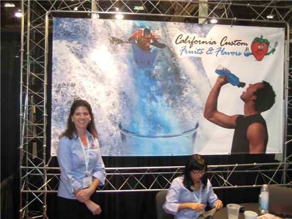 A woman in a light blue shirt stands beside a seated woman at a table, under a California Custom Fruits & Flavors banner showing a kayaker and a man drinking from a bottle.